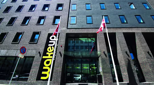 Eksteriør av en moderne murbygning med rektangulære vinduer. Et stort gult og svart "Wakeup"-skilt vises nær inngangen, flankert av to flaggstenger med røde og hvite flagg. Himmelen er klar og blå.