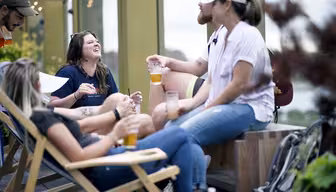 En gruppe mennesker slapper av på en takterrasse, sitter på stoler og benker og nyter drinker. De ler og står overfor hverandre. Innstillingen er uformell med grøntområder i bakgrunnen.