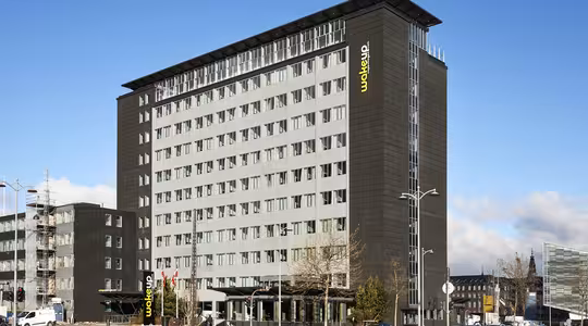 En moderne hotellbygning i flere etasjer med en elegant svart fasade og store vinduer. Hotellets navn vises vertikalt i øvre hjørne. Forgrunnen har biler, trær og en klar blå himmel.
