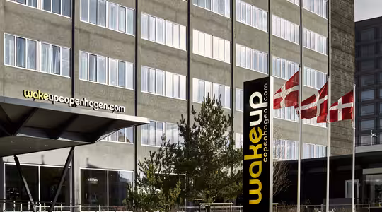 Moderne hotellbygg med store vinduer og skiltet "Wakeup Copenhagen" synlig. Tre danske flagg vaier på stenger utenfor inngangen. Noen få små trær er foran bygningen.