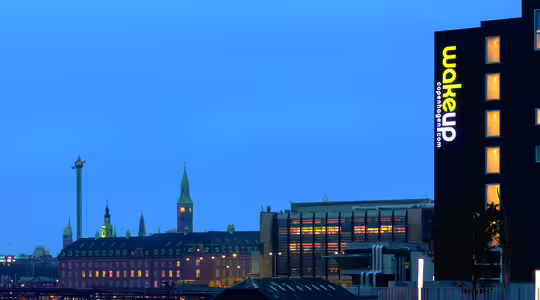 Skyline i skumringen med bygninger og et sterkt opplyst «wakeup copenhagen»-skilt på en mørk bygning til høyre. Et tårn med skyss er synlig til venstre. Himmelen er myk blå, og bygningene er opplyst, noe som gir varme til scenen.