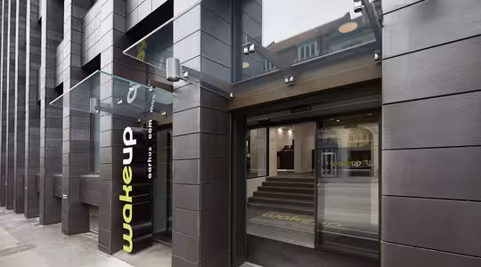 Utvendig visning av en moderne bygning med mørkt panel, med en baldakin i glass over en inngang. Inngangspartiet har store skyvedører i glass med teksten «wakeup copenhagen» på. Innvendig er det trappetrinn som fører opp til hovedområdet.