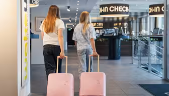 To personer med langt hår, sett bakfra, går med rosa rullekofferter mot en hotellresepsjon. De er uformelt kledd, den ene i en hvit skjorte og jeans, den andre i en tie-dye skjorte. Skilt for innsjekking er synlig ovenfor.
