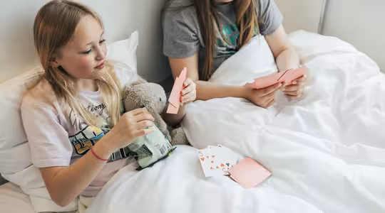 To barn sitter på en seng med hvite puter og tepper og spiller kort. Det ene barnet holder en bamse og en snackpose, det andre holder spillekort. Et lite vindu kan sees i bakgrunnen.