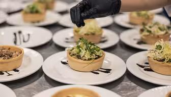 Nærbilde av en kokkes hånd iført en svart hanske, pyntet flere tallerkener med velsmakende terter fylt med grønt og en skvett mørk saus på hvite tallerkener, pent arrangert på en overflate i rustfritt stål.