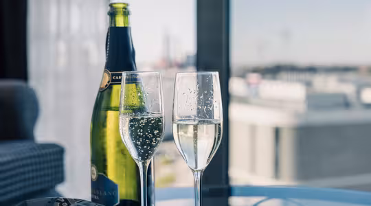 To fylte champagnefløyter står på et glassbord ved siden av en åpen champagneflaske. Bakgrunnen har et bybilde sett gjennom store vinduer, med en sofa delvis synlig til venstre.