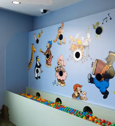 Et fargerikt lekeromshjørne med en lyseblå vegg med tegneseriedyr som spiller musikkinstrumenter. Nedenfor ligger en ballgrop med flerfargede baller mot veggen, og legger til en leken atmosfære.