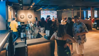 En moderne bar med sosialt samvær. En kvinne i en svart topp står i forgrunnen nær disken, med andre samlet rundt bord og spiller dart i bakgrunnen. Belysningen er varm, og skaper en koselig atmosfære.