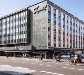 En moderne bygning i flere etasjer med et stort skilt som viser Imperial. Fasaden inkluderer mange vinduer. Det er en restaurant på gateplan med uteservering, og folk går og sykler i nærheten på en solrik dag.