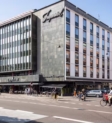 En moderne bygning i flere etasjer med et stort skilt som viser Imperial. Fasaden inkluderer mange vinduer. Det er en restaurant på gateplan med uteservering, og folk går og sykler i nærheten på en solrik dag.