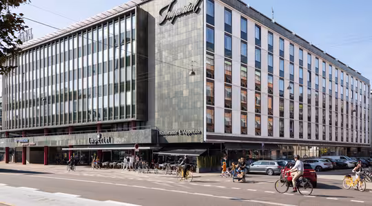 En moderne bygning i flere etasjer med et stort skilt som viser Imperial. Fasaden inkluderer mange vinduer. Det er en restaurant på gateplan med uteservering, og folk går og sykler i nærheten på en solrik dag.
