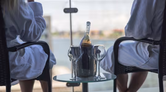 To personer i badekåper sitter på en balkong med glassrekkverk med utsikt over en vannmasse. En flaske champagne i en isbøtte og to glass står på et lite rundt bord mellom dem.