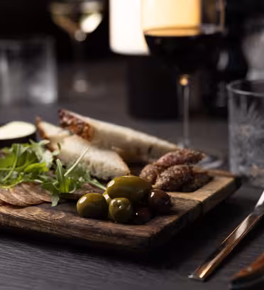 En treplate med assortert charcuterie: oppskåret kjøtt, oliven, ost og brød. Friske grønnsaker pynter fatet. I nærheten står et glass rødvin og et krystallglass på det mørke trebordet, dekket med kniv og gaffel og en sammenrullet serviett.