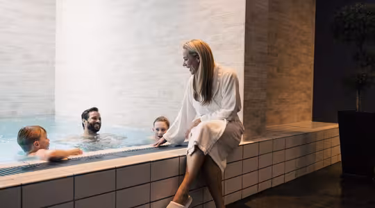 En kvinne i hvit badekåpe og tøfler sitter ved kanten av et innendørsbasseng og smiler til tre personer som er i vannet. Scenen er varm og avslappende, med moderne fliser og dempet belysning som forsterker den koselige atmosfæren.