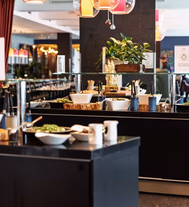 Buffetoppsett i en restaurant med en rekke retter i hvite skåler på mørke benkeplater. Fargerike pendellamper og planter legger til dekor. Spisebord med stoler er synlige i bakgrunnen, og skaper en innbydende atmosfære.