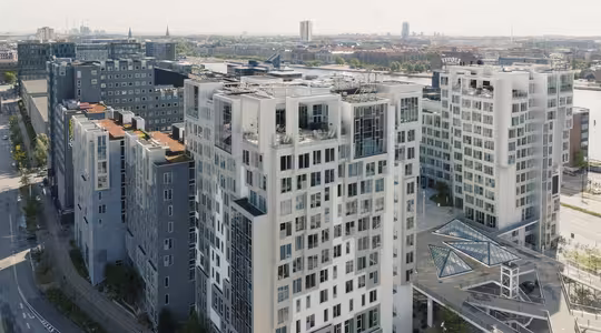 Luftfoto av moderne høyhus i et urbant område. Arkitekturen har en kombinasjon av glass og betong med geometriske design. I bakgrunnen er en elv og bybilde synlig under en overskyet himmel.