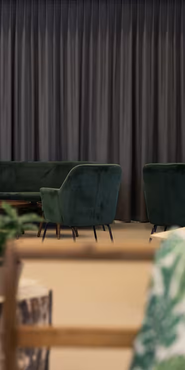 En koselig sittegruppe med grønne fløyelsofaer og -stoler, et salongbord i tre og innredning med plantetema. Bakteppet har gulvlange grå gardiner og en elegant, svart salgsautomat med "Tivoli"-grafikk. Naturlig belysning forbedrer plassen.