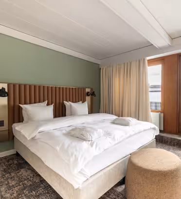 Et moderne hotellrom har en stor seng med hvitt sengetøy og en hodegjerde i tre mot en salviegrønn vegg. Det er en beige skammel ved foten av sengen, og store vinduer med tunge gardiner vender ut mot et industrielt bybilde.