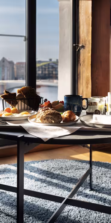 Et frokostbrett med brød, bakverk, egg og drikke står på et rundt bord ved store vinduer med utsikt over et bybilde ved vannkanten. Sollys fyller det moderne rommet med et teppe og en oransje stol.