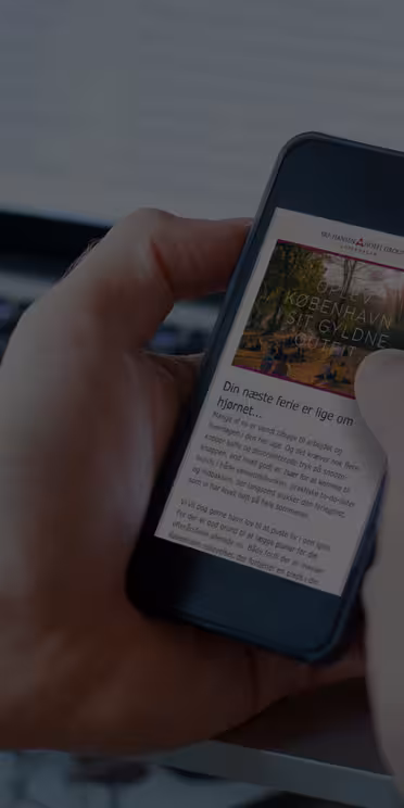 En person holder en smarttelefon som viser en nettside med tittelen "Norsk Havarnas Oljeind" foran en bærbar datamaskin. Bakgrunnen er litt uskarp, og personen bruker begge hendene, med en tommel som ruller på telefonens skjerm.