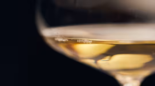 Nærbilde av et delvis fylt vinglass med en gylden væske, sannsynligvis hvitvin. Bakgrunnen er mørk, og skaper en kontrast som fremhever klarheten og fargen til vinen.