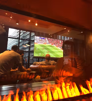Folk som ser på en fotballkamp på en stor TV i et moderne rom med gulv-til-tak-vinduer. Scenen sees gjennom et glasspanel som reflekterer flammer fra en nærliggende peis, og skaper en varm atmosfære.