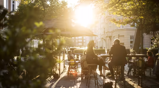 Folk sitter ved utendørs kafébord under parasoller og nyter en solrik dag. Sollyset filtrerer gjennom trærne og kaster flekkete skygger på bakken. Scenen formidler en avslappet og varm atmosfære.