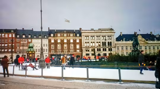Folk skøyter på en skøytebane i urbane omgivelser, omgitt av historiske europeiske bygninger. Scenen inkluderer noen få tilskuere og nærliggende statuer, med en overskyet himmel over hodet.