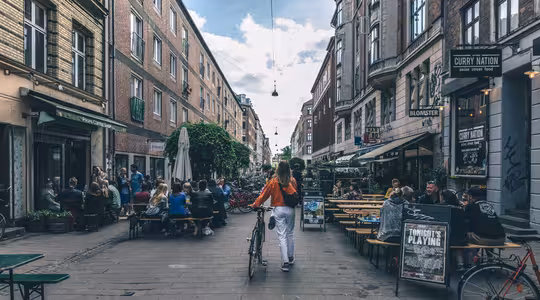 En kvinne i rød jakke går på sykkel nedover en livlig gate med utekafeer og restauranter. Folk sitter ved bordene på begge sider og nyter en solrik dag. Gaten er omgitt av murbygninger under en delvis overskyet himmel.