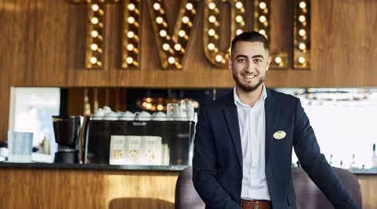 En mann i dress står smilende foran en trevegg med «TIVOLI» i opplyste bokstaver. Bak ham står en disk med kaffeutstyr og bøker.