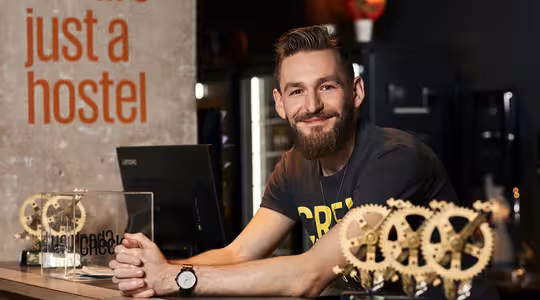 En mann med skjegg står og smiler bak en disk og viser frem utstyr og en prisplakett. Bak ham er det en vegg med ordene "vi er bare et herberge" i stor tekst.