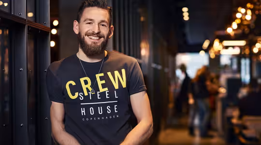 En skjeggete mann iført en T-skjorte med teksten «CREW STEEL HOUSE COPENHAGEN» lener seg mot en vegg i en svakt opplyst korridor. Han smiler med armene i kors. Bakgrunnen har varme lys og uskarpe figurer.