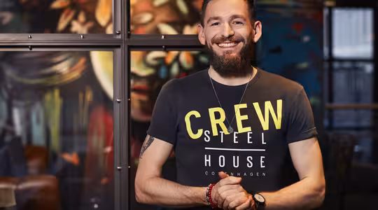 En smilende mann med skjegg står foran en fargerik, kunstnerisk bakgrunn. Han har på seg en svart t-skjorte med ordene «CREW STEEL HOUSE COPENHAGEN» og har hendene sammen. Rommet har en moderne, kreativ atmosfære.