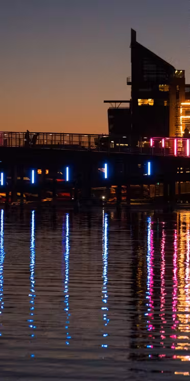 Nattutsikt over et bybilde med en bro med fargerike neonlys i rosa, blått og grønt. Lysene reflekterer levende på vannet under. I bakgrunnen står opplyste moderne bygninger mot kveldshimmelen.