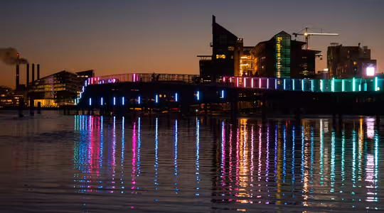 Nattutsikt over et bybilde med en bro med fargerike neonlys i rosa, blått og grønt. Lysene reflekterer levende på vannet under. I bakgrunnen står opplyste moderne bygninger mot kveldshimmelen.