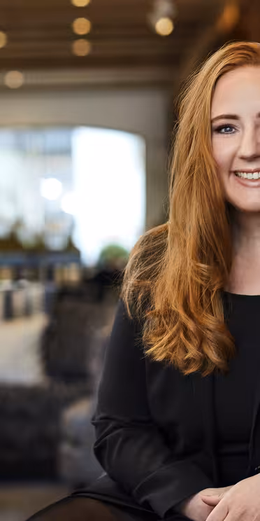 En person med langt rødt hår, iført svart blazer og navneskilt, sitter innendørs. Bakgrunnen har et moderne, godt opplyst salongområde med dempet belysning, puter og stearinlys på et bord.