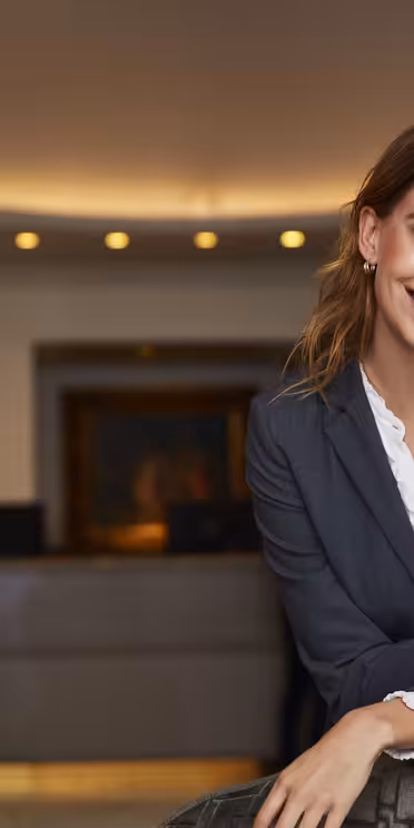 En person i profesjonell dress smiler mens han lener seg på en stol i en hotellobby. Lobbyen har en varmt opplyst resepsjon i bakgrunnen med en medarbeider som tar seg av en gjest. Atmosfæren er innbydende og elegant.