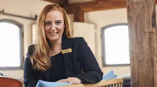 En person med langt rødt hår og navneskilt smiler, sittende på en stol med blå pute, i et varmt opplyst rom med trebjelker og buede vinduer. De har på seg en svart blazer.