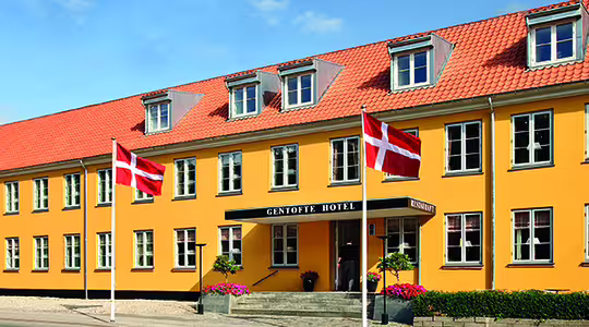 En knallgul bygning med rødt tak, med et skilt med teksten "GENTOFTE HOTEL." To danske flagg vises foran. Inngangen har trappetrinn, potteplanter og en liten baldakin. Klarblå himmel over hodet.