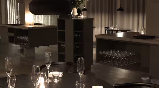 Svakt opplyst restaurantinteriør med moderne innredning. Sirkulære bord er dekket med vinglass og servietter. Et stort svart pendellys henger over. Hyller i bakgrunnen viser flere glass, og en vase med blomster pryder rommet.