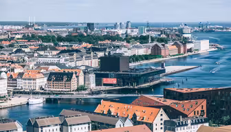 Utsikt over et bybilde ved vannet, med en blanding av historisk og moderne arkitektur. Bygninger med røde tak langs havnen, med båter i vannet. En moderne glassbygning skiller seg ut, med en fjern utsikt over en bro og kraner i horisonten under en delvis overskyet himmel.