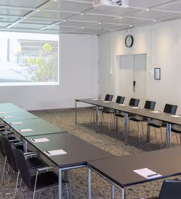 Et moderne konferanserom satt opp med to lange U-formede bord, svarte stoler og en projeksjonsskjerm som viser et bilde. Rommet har et nøytralt teppe, veggklokke og skarp belysning.