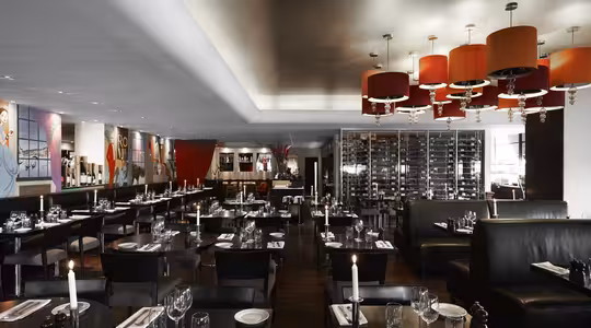 Elegant restaurantinteriør med dempet belysning, med moderne svarte bord dekket med glass og hvite servietter. Røde lysekroner henger i taket, og et stort vinstativ og fargerike veggmalerier pryder veggene.
