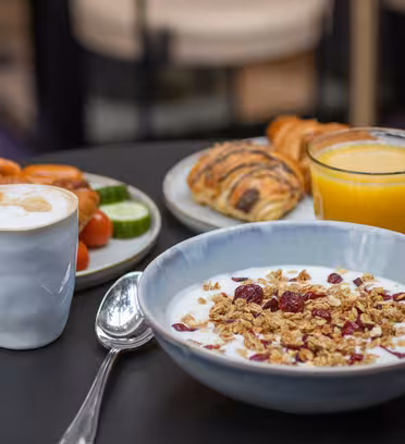 Et frokostpålegg med en bolle med yoghurt toppet med granola og tørkede bær, en kopp cappuccino, et glass appelsinjuice og en tallerken med bakverk, agurker, tomater og appelsinskiver på et svart bord.