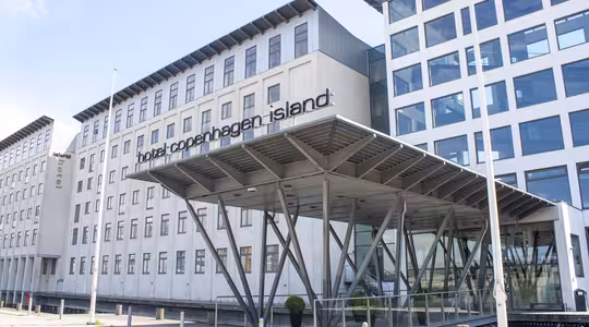 Moderne hotellbygning med baldakininngang støttet av metallbjelker, med store vinduer og et skilt med teksten "Hotel copenhagen island". Bygget er i flere etasjer med en blanding av designelementer i glass og betong.