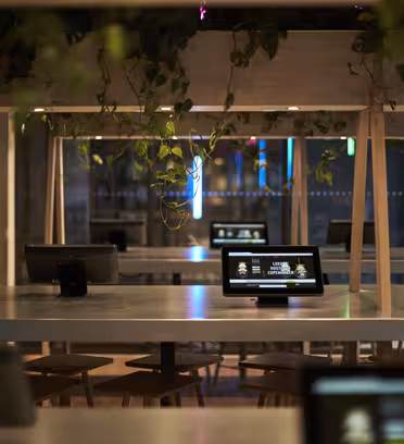 En moderne, svakt opplyst kafé med langbord og nettbrett med berøringsskjerm på. Hengende planter dekorerer rommet, og et stort vindu reflekterer neonlys utenfra. Oppsettet kombinerer teknologi med en koselig atmosfære.