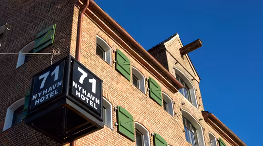 En murbygning med grønne vindusskodder og et skilt med "71 Nyhavn Hotel" mot en klarblå himmel.