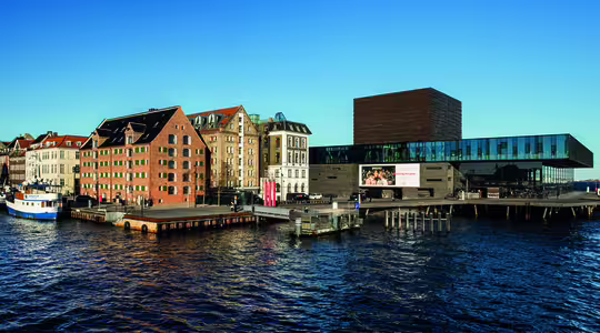 Utsikt ved vannet av et bybilde med en rekke fargerike, historiske bygninger langs en kanal. En moderne glass- og mørkt trestruktur er fremtredende plassert på vannet, ved siden av forankrede båter under en klarblå himmel.