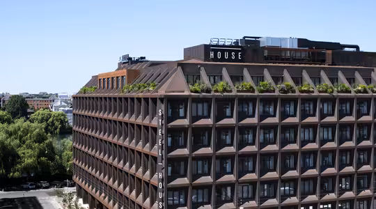 En stor bygning i industristil med en rutenettlignende fasade og takgrønt, merket «Stålhus». Det er satt mot en klar blå himmel, med omkringliggende trær og fjern arkitektur synlig.