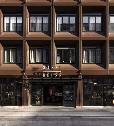 Bildet viser en moderne bygning med en rutenettlignende fasade med store vinduer. Inngangen viser navnet "Stålhus" med hvite bokstaver over glassdører. Bygget har en brun, industriell estetikk med noe grønt rundt inngangspartiet.